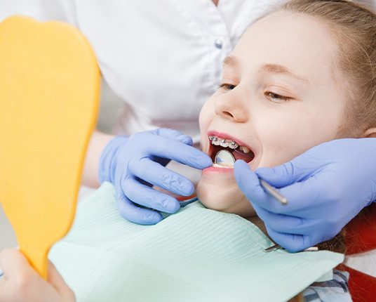 a child with braces looking in a handheld mirror while a dental hygienist holds a mirror in their mouth