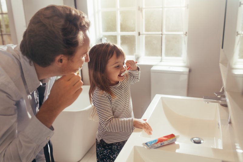 Father brushing his teeth watching his young daughter brush hers at a bathroom sink